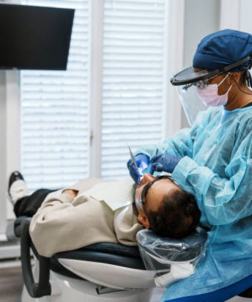 Multiracial, female patient sits, performing a tooth filling procedure on a male patient of Indian descent.