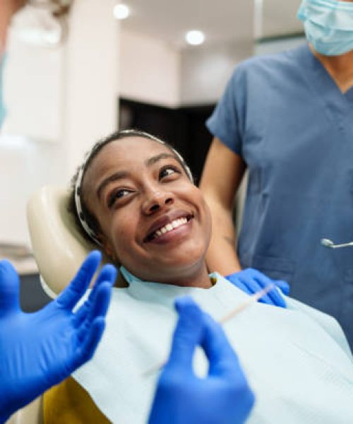 A Black woman smiles warmly while sitting in a dental chair. She is attended by a diverse dental team, including a male and a female, both wearing blue scrubs and gloves. The setting is bright and modern.